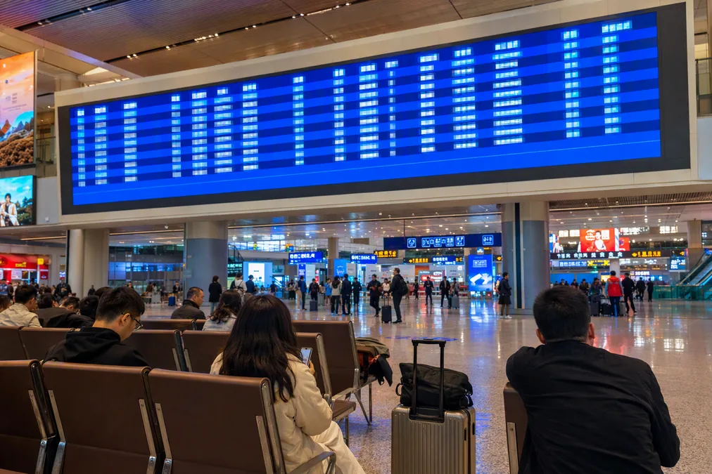 A large departure board above the waiting hall of a Chinese high-speed railway station, with passengers seated below